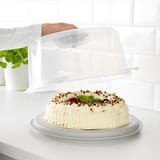 A cake in a carrier with a clear lid, placed on a countertop. The carrier has grooves and smooth sides for different pastries.