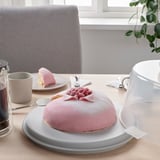 Cake in carrier with lid and handle, shown on table with plate and cups.