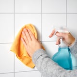 Person cleaning tiles with yellow cloth, holding blue spray bottle.
