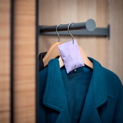 A teal coat hangs on a closet rod, a lavender-scented sachet labeled KROPPSVARM attached, releasing fresh scent.
