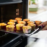 Hand holding tray of golden muffins in colorful liners being removed from oven.