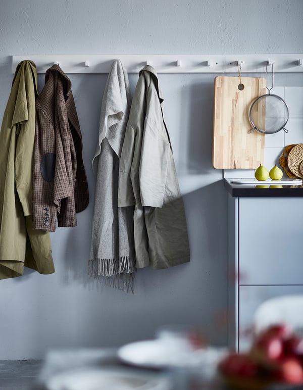 White KUBBIS coat rack with hooks, scarves, jackets. Kitchen counter with cutting board, strainer, fruits.