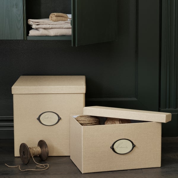 Two KVARNVIK beige storage boxes, one closed, one open, on dark floor, with woven texture, label holders, dark green cabinet background.