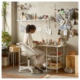 A woman works at a white LAGKAPTEN desk with shelves, surrounded by pottery and plants, with a bulletin board behind her.