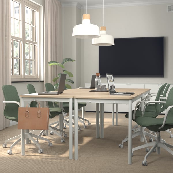Modern office with LÅNGFJÄLL chairs, wooden table, two pendant lamps, mounted tv, window, green chair with bag, laptop, and plant.