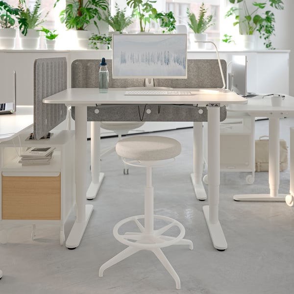 Modern office setup with white LIDKULLEN stool, adjustable height desks, grey dividers, and multiple potted plants for decoration.