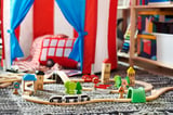 Colorful wooden toy train track with green trees, blue tunnel, and small buildings on a carpeted floor.