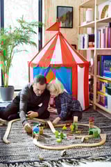 A man and child play with wooden LILLABO train set, red and white tent, bookshelves and potted plant in room.