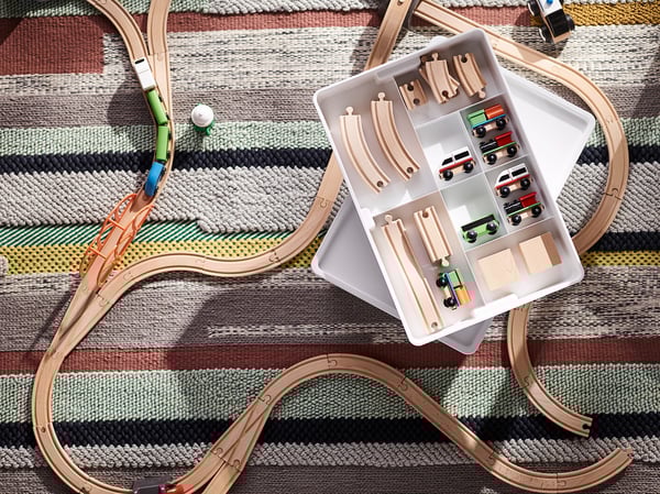 Wooden train set with tracks, cars, and storage box on a striped rug.