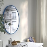 An oval mirror with a blue frame on a wall, reflecting a building exterior, placed above a white table with a lamp and decor items.