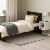 Modern bedroom with black bed, white pillows, grey blanket, grey rug on wooden floor, small plants, and white nightstand.