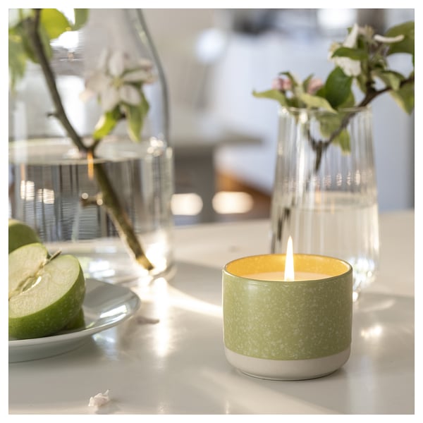 LÖVSKOGSLUND candle in green, ceramic, sits on a table with apples, flowers, lighted.