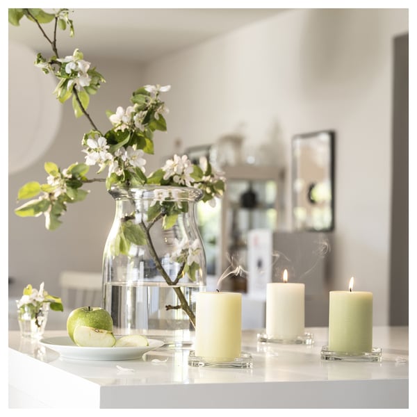 Three lit beige candles on glass holders, a clear vase with green apples and white flowers, on a white surface.