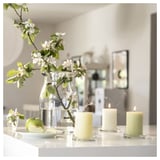 Three lit beige candles on glass holders, a clear vase with green apples and white flowers, on a white surface.