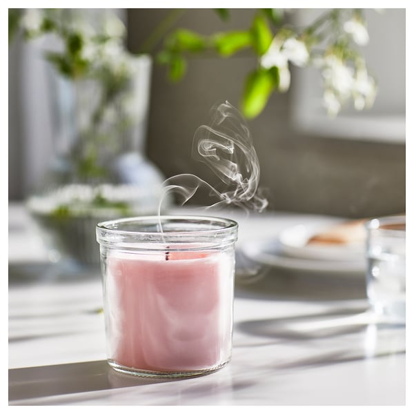 A burning pink scented candle in a clear glass jar on a light-colored table. Soft smoke curls upwards, surrounded by green plants and natural light.