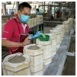 A man wearing a red shirt and face mask works in a ceramic factory, painting earthy tone vases by hand.