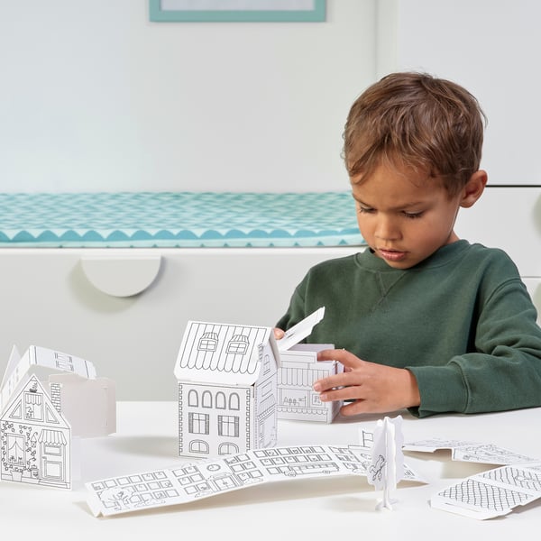 A young boy is crafting with the MÅLA cardboard house templates, developing fine motor skills and creativity by assembling and coloring a small town.