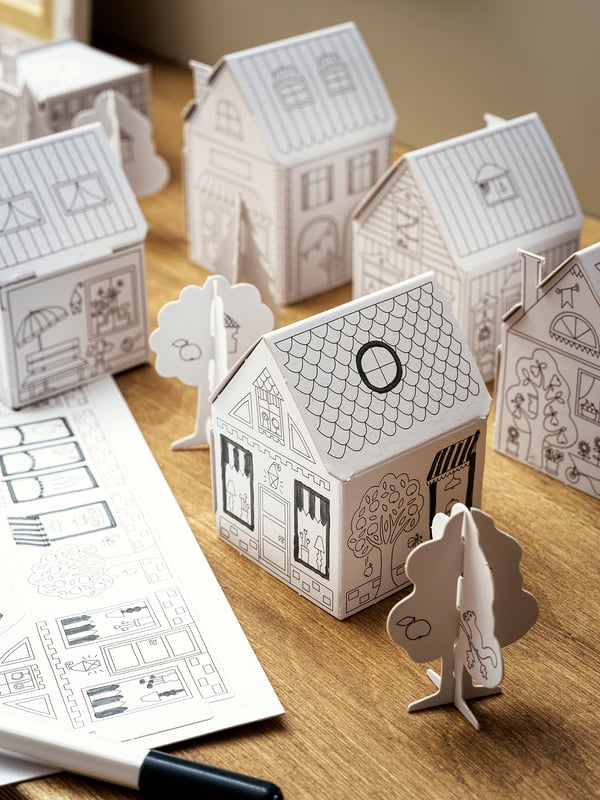 Black and white foldable cardboard houses and trees sit on a wooden table, designed for children to paint and create a small town.