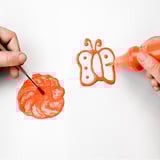 Childs hands painting butterfly with orange brush and squeezing paint from bottle on white surface.