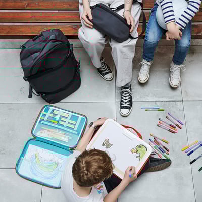 Children on bench, one drawing with open MÅLA art case. Colorful supplies and artwork on ground, large backpack beside them.