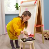 Young girl wearing a bright yellow apron stands at an easel, mixing paints in a palette. She is focused on her artwork, with a teddy bear beside her.