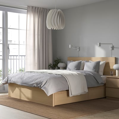 Modern bedroom with light wood bed, grey linens, pendant lamp, tall curtain, small plant, wooden nightstands, wall lamps, and minimalistic design.