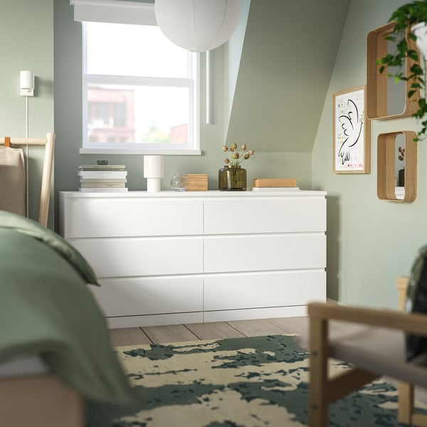 MALM chest against green wall with books, lamp, vase. Tall, matches nearby chairs. Park print right, window above.