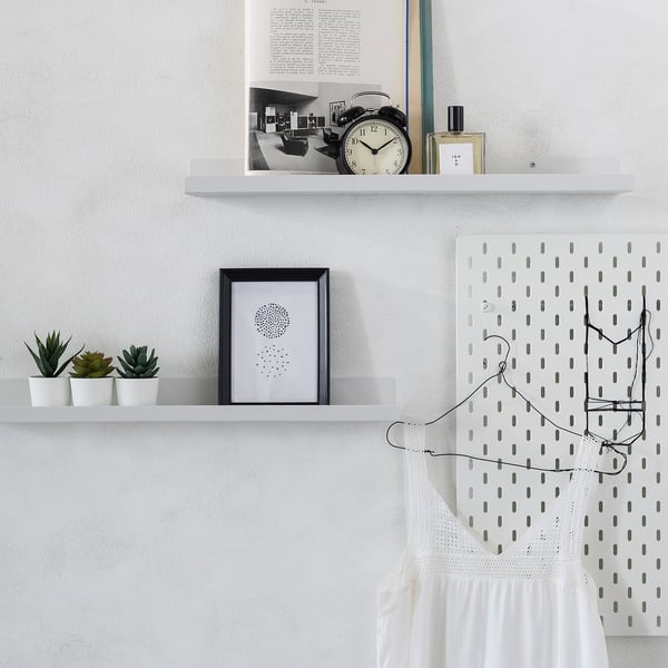 Three white shelves with black accents hold items like a clock, plants, and art. A pegboard with hanging lights and a white dress is beside it.