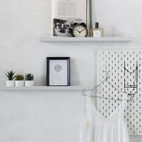 Three white shelves with black accents hold items like a clock, plants, and art. A pegboard with hanging lights and a white dress is beside it.