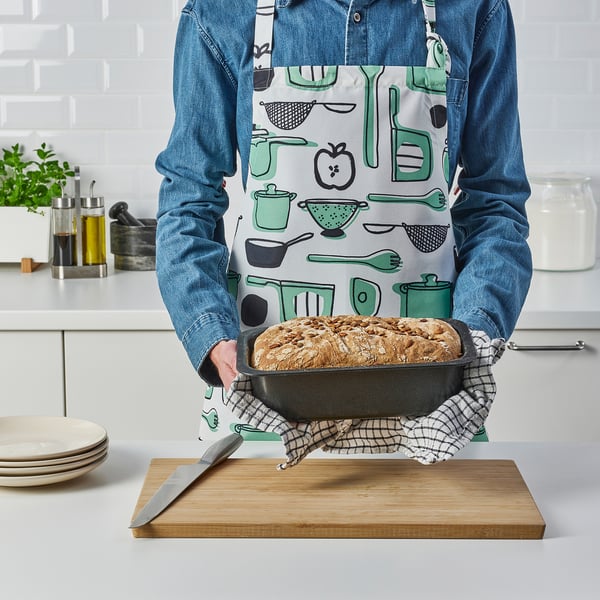 Person holding a freshly baked bread in a loaf tin on a kitchen counter. The apron has a cooking tools print.