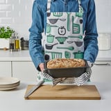 Person holding a freshly baked bread in a loaf tin on a kitchen counter. The apron has a cooking tools print.