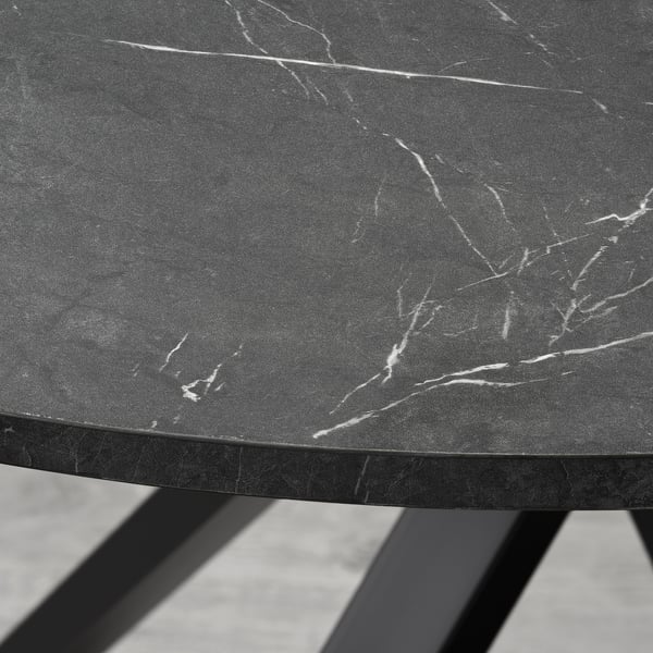 A close-up of a modern, round tabletop with a sleek, dark finish and subtle white veining patterns, supported by black metal legs.