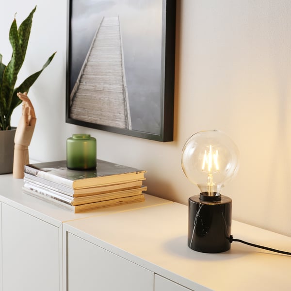 MARKFROST table lamp with black marble base and clear bulb on storage unit, beside books, green vase, and plant.