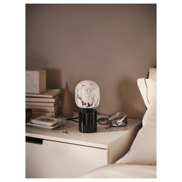 Modern bedroom with white bed, nightstand stocks books, & white marble lamp.