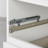 Close-up of soft-closing drawer mechanism; shows metallic slide, gray damper, and roller wheel on side of white cabinet.