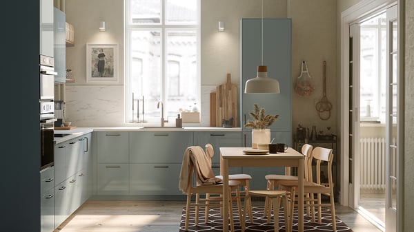 Modern kitchen with grey-blue METOD cabinets, wooden table, and chairs. Natural light from large window, pendant light, and wall art.