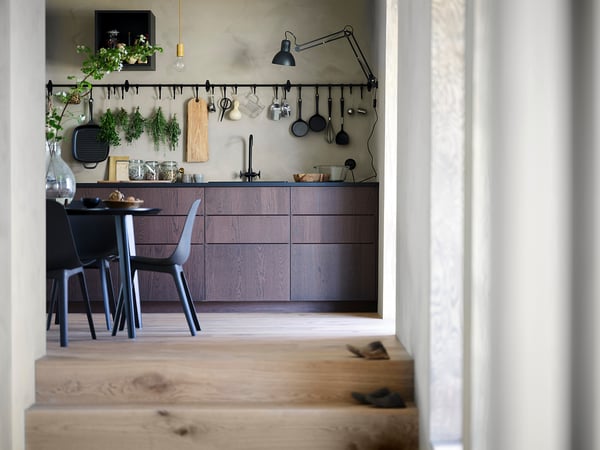 Modern kitchen with dark brown cabinets, light wood floors, hanging plants, black chairs, industrial lamp, and hanging utensils.