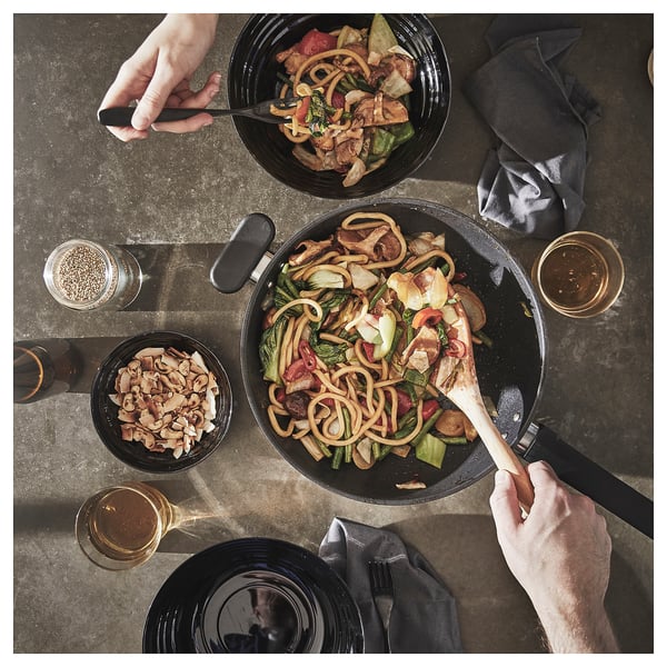 Two people cooking noodles in black wok; one stirs, other serves.
