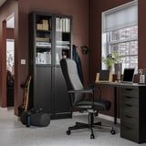 Modern home office setup. Black MILLBERGET chair, desk with laptop, bookshelf with books, skis, and headphones.