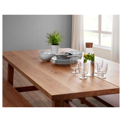 Displays a MÖCKELBY wooden dining table with a bench and decorative items, including plates, glasses, and a plant.