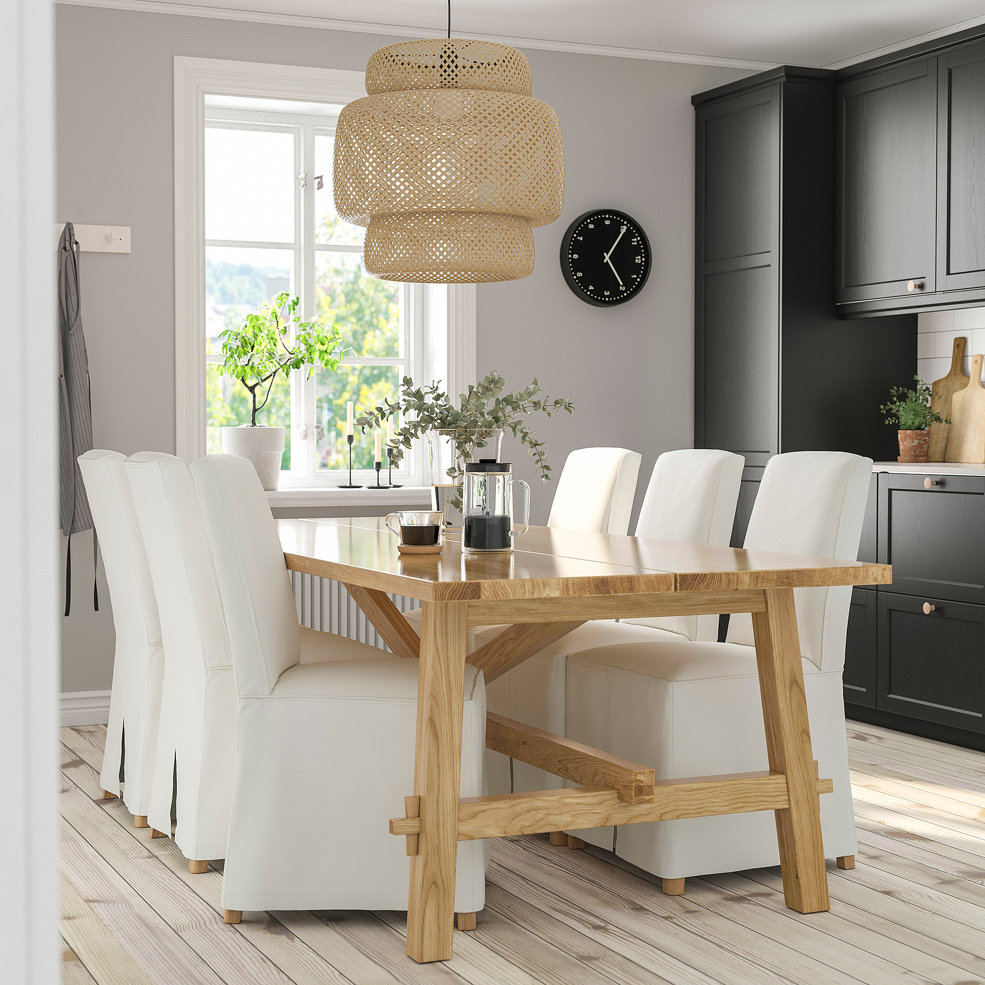 Modern dining area with light wood table, six white chairs, wicker pendant lamp, and wall clock. Kitchen visible in background.