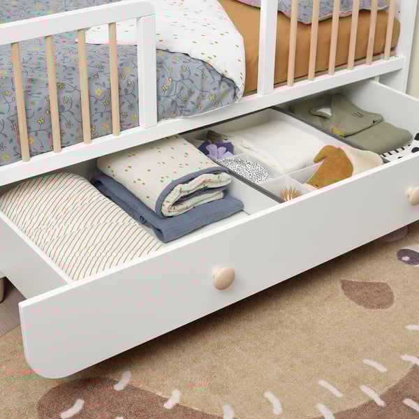 White kids bed with adjustable, open drawer, neatly folded blankets. Floral and striped patterns visible, showing storage use.