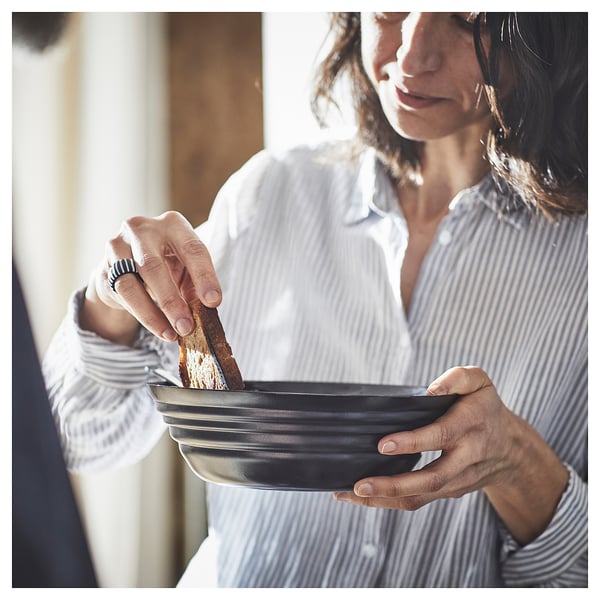 A woman wearing a striped shirt holds a black, round bowl, dipping a piece of bread.