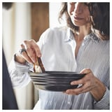 A woman wearing a striped shirt holds a black, round bowl, dipping a piece of bread.