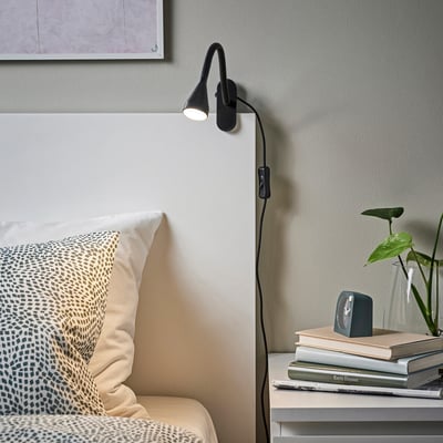Black NÄVLINGE lamp on white headboard, lighting nightstand with books & plant. Patterned pillow on bed.