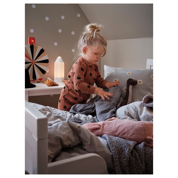 A child playing on a bed with a daisy-patterned pillow. The room has fairy lights, a night lamp, and a colorful spinning top on a pink shelf.