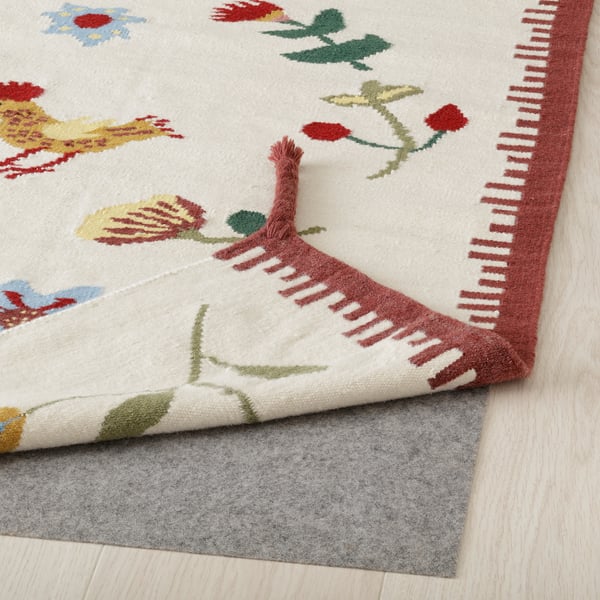 Decorative rug with vibrant floral patterns and red braided corners laid on a wooden floor with gray felt underlay visible at one corner.