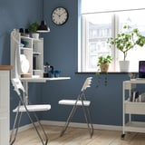 Compact workspace: white drop-leaf table with storage, two folding chairs, plant cart, and window view.