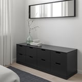 Black NORDLI 6-drawer chest in bedroom, mirror, window with sheer curtains, vase with branches.