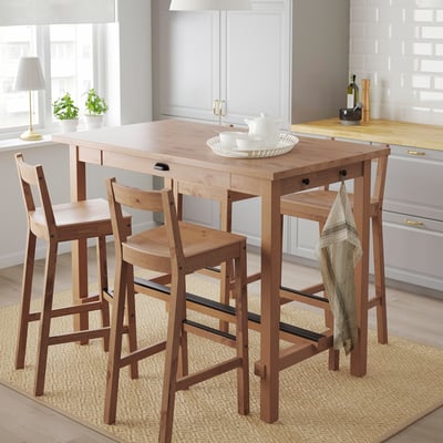 Small kitchen with light-colored wooden NORDVIKEN bar set. Three stools by wooden table, tea set on table. Bright, airy room with plants.